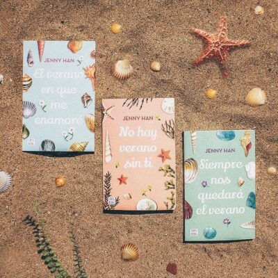 Tres libros de la trilogía "Verano" sobre una playa arenosa, con caracolas marinas decorativas en la carátula.